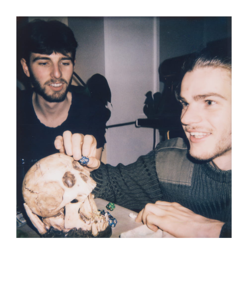 Two friends playing D&D at a table with dice and a human skull prop, engrossed in conversation during their role-playing game.