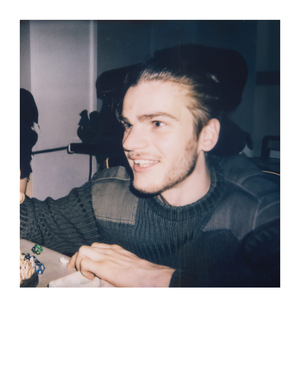 Young man with light-colored hair, smiling and leaning on a table with dice and other items, in a warmly lit room.