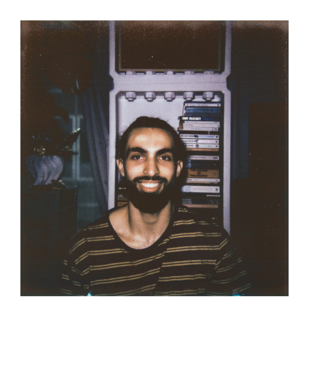 Smiling man with a striped shirt, seated in front of a shelf stacked with books and a fridge in the background.