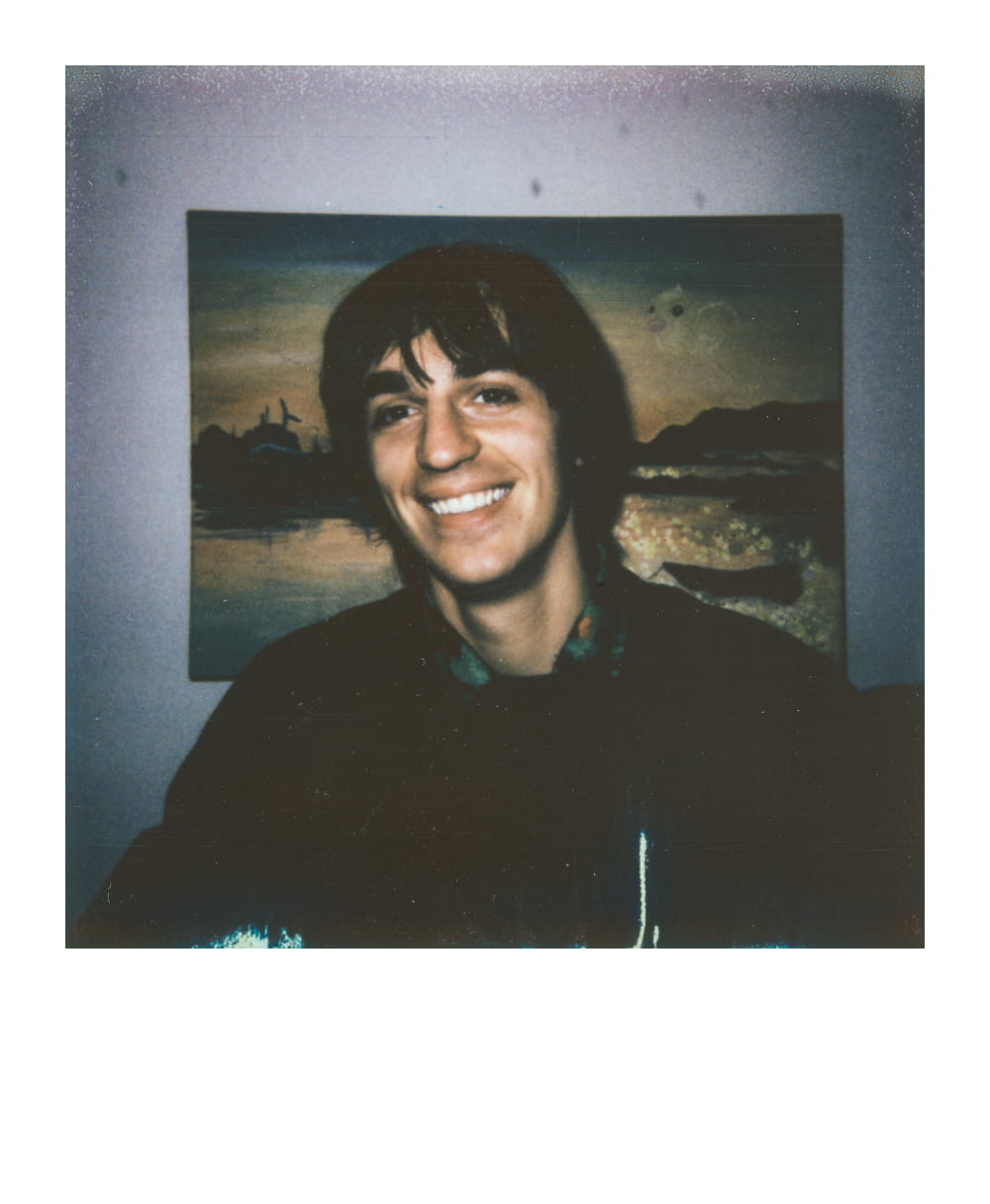 Man with shaggy hair and a big smile, sitting in front of a painting depicting a sunset over water.