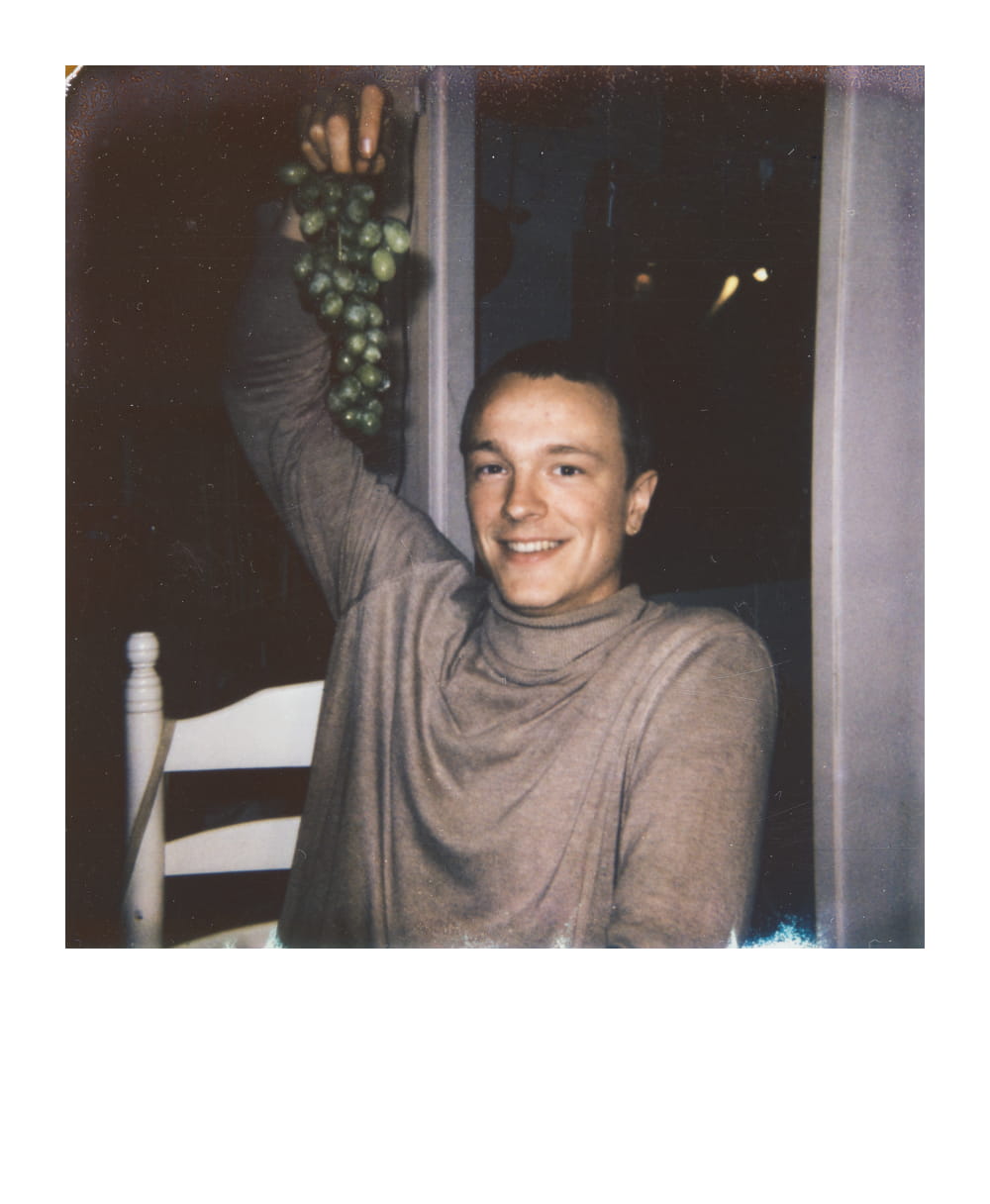 Dungeon Master with light hair, smiling while sitting at a table with a vine of grapes in one hand.